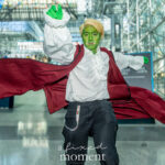 Cosplayer @pasionplays dressed as an orc adventurer with green skin, blonde hair, and tusks lunges forward dramatically in a burgundy coat and white ruffled shirt at New York Comic Con 2025 inside the Javits Center.
