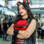Cosplayer @antiv0gue dressed in a red and black devil-inspired costume with sparkling horns, red eyeshadow, and a confident pose at New York Comic Con 2025 inside the Javits Center.