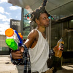Cosplayer @thee_evantures_of_cosplay dressed as Ashtray from Don’t Be a Menace to South Central While Drinking Your Juice in the Hood, posing outside the Javits Center at New York Comic Con 2025, holding a toy water gun and bottle, wearing a white tank top, plaid shirt, tan pants, and a tall braided hairstyle with colorful beads.
