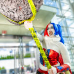 Cosplayer @hell.baby.13 dressed as Harley Quinn, wearing a red and blue metallic costume and holding a giant mace wrapped in yellow caution tape, posing energetically inside the Javits Center at New York Comic Con 2025.