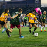 Soccer players in yellow and green jerseys chase the ball during “The Cost of Living Classic” at Maimonides Park in Coney Island, part of Zohran Mamdani’s campaign event on October 19, 2025.