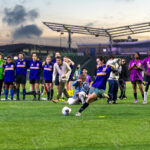 A player in a blue “Brooklyn” jersey kicks a soccer ball during a penalty shot as the Bronx team watches at “The Cost of Living Classic” soccer tournament in Coney Island.