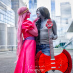 Cosplayers @xjesswasiakx and @girlbug000027 dressed as Princess Bubblegum and Marceline from Adventure Time, smiling affectionately while holding a red axe-shaped bass guitar outside the Javits Center at New York Comic Con 2025.