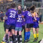 A group of soccer players in purple jerseys that read “For a city we can afford” celebrate together on the field at Maimonides Park during “The Cost of Living Classic” in Coney Island.