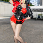 Cosplayer @miss_sam_mae dressed as Harley Quinn in a red and black latex outfit with a hooded jacket and red sneakers, striking a playful pose outside the Javits Center at New York Comic Con 2025 in NYC.