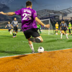 A soccer player wearing a purple Queens jersey with the slogan “for a city we can AFFORD” takes a corner kick under the stadium lights during “The Cost of Living Classic” at Maimonides Park in Coney Island.