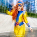 Cosplayer @amaliaaacat dressed as Jean Grey from X-Men in a yellow and blue bodysuit with a red X belt and red hair, posing outside the Javits Center at New York Comic Con 2025 in NYC.