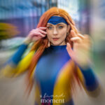 Cosplayer @amaliaaacat dressed as Jean Grey from X-Men, wearing a blue and yellow bodysuit and red wig, posing with hands raised in a telekinetic stance outside the Javits Center at New York Comic Con 2025.