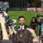 Zohran Mamdani smiles as reporters and photographers crowd around him for interviews after “The Cost of Living Classic” soccer event at Maimonides Park in Coney Island.