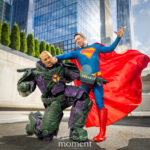 Cosplayers @dannycarmelo as Superman and @jamal_johns as Lex Luthor reenacting a battle scene, with Superman punching Lex Luthor in his green-and-purple power armor outside the Javits Center at New York Comic Con 2025.