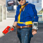 Cosplayer @insomniacwebslinger dressed as a Vault Dweller from Fallout, wearing a blue and yellow vault suit, sunglasses, and a Pip-Boy on his arm, holding a red Nuka-Cola blaster outside the Javits Center at New York Comic Con 2025.