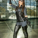 Cosplayer @racksonrays dressed as Catwoman from DC Comics, posing with a black whip over her shoulder, wearing thigh-high leather boots, a corset, and a cat-ear headpiece at New York Comic Con 2025 inside the Javits Center.