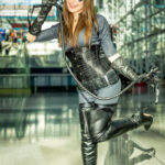 Cosplayer @racksonrays dressed as Catwoman from DC Comics, posing playfully with one leg raised and holding a whip while smiling inside the glass atrium of the Javits Center during New York Comic Con 2025.