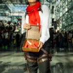 Cosplayer @detrmik dressed as Mikasa Ackerman from Attack on Titan, wearing a white shirt, red scarf, and brown harness straps, holding an open wooden box with a syringe prop at New York Comic Con 2025 inside the Javits Center.