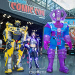 Three cosplayers at New York Comic Con 2025: Bumblebee from Transformers, a female cyberpunk-style cosplayer with glowing purple LED armor, and Galactus from Marvel Comics, standing together in front of the New York Comic Con banner at the Javits Center.