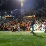 A large group of players and fans pose together on the field under bright stadium lights at Maimonides Park during “The Cost of Living Classic” soccer tournament in Coney Island.