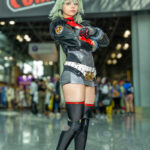 Cosplayer @xenluvsmiku dressed as Silver Wolf from Honkai: Star Rail, standing confidently with arms crossed in front of the New York Comic Con banner at the Javits Center, wearing a silver and black outfit with red accents and mint green hair.