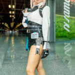 Cosplayer @hharthere dressed as Mash Kyrielight from Fate/Grand Order, wearing a white blouse, black harness vest, skirt, gloves, and glasses with pink hair, posing confidently inside the Javits Center at New York Comic Con 2025.