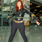 Cosplayer @a_l_a_n_is dressed as Marvel’s Black Widow at New York Comic Con 2025, wearing a black tactical suit with gold accessories and holding batons in a power pose inside the Javits Center.