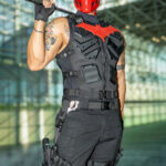 Cosplayer @ryfam_ dressed as Red Hood from DC Comics, wearing a black tactical vest with a red bat emblem, red mask, and combat pants, posing with a crowbar inside the Javits Center at New York Comic Con 2025.