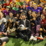 Zohran Mamdani, dressed in a black suit, smiles while sitting on the field surrounded by players in colorful jerseys from all borough teams after “The Cost of Living Classic” at Maimonides Park in Coney Island.
