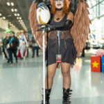 Cosplayer @chunslii34 dressed as Lilith Clawthorne from The Owl House at New York Comic Con 2025, wearing brown and black feathered wings, a dark brown outfit, black gloves, and holding a staff topped with an owl palisman inside the Javits Center.