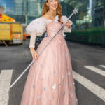 Cosplayer @mchalelv dressed as Glinda the Good Witch from The Wizard of Oz at New York Comic Con 2025, wearing a pink sparkling gown, tall jeweled crown, and holding a silver wand with a star tip outside the Javits Center in New York City.
