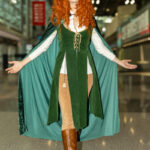 Cosplayer @maker_baker_picturetaker dressed as an elven sorceress in a green velvet vest, cloak, and brown boots, walking with arms open inside the Javits Center at New York Comic Con 2025 in NYC.