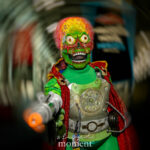 Cosplayer @bombshellbeckie dressed as a Martian invader from Mars Attacks! at New York Comic Con 2025, wearing a green bodysuit, metallic chest armor, and red cape, holding a silver ray gun and posing inside the Javits Center.