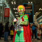 Cosplayer @bombshellbeckie dressed as a Martian invader from Mars Attacks! at New York Comic Con 2025, wearing a green bodysuit, metallic chest armor, and red cape, holding a silver ray gun and posing inside the Javits Center.