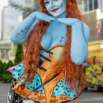 Cosplayer @chaosplaykarma dressed as Sally from The Nightmare Before Christmas with blue body paint, long red hair, and a colorful patchwork dress, posing outside the Javits Center during New York Comic Con 2025.