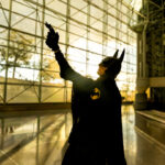 Cosplayer @bloodbbyblood dressed as Batman at New York Comic Con 2025, standing in silhouette inside the Javits Center with sunlight streaming through the glass windows, pointing a grappling gun upward.