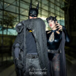 Cosplayer @bloodbbyblood as Batman poses with a Catwoman cosplayer inside the Javits Center at New York Comic Con 2025. Batman wears a black distressed cape and cowl, while Catwoman is dressed in a glossy black bodysuit with goggles and tattoos visible on her legs.