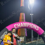 Zohran Mamdani speaks on stage beneath a pink “CHAMPIONS” arch at Maimonides Park in Coney Island, with the illuminated Parachute Jump tower glowing behind him.