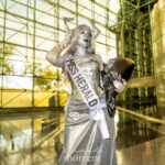 Cosplayer @kailcarps dressed as Miss Herald at New York Comic Con 2025, wearing a shimmering silver gown, white “MISS HERALD” sash, silver horns, satin gloves, and holding a black-wrapped bouquet, posing under sunlight inside the Javits Center.