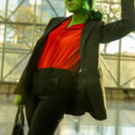 Cosplayer @_lao_you_tiao dressed as She-Hulk: Attorney at Law at New York Comic Con 2025, wearing a red blouse and black blazer with green body paint, flexing her arm confidently inside the Javits Center.