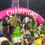 Zohran Mamdani celebrates with the Brooklyn soccer team as they hold a golden trophy and champagne under a pink “CHAMPIONS” arch at Maimonides Park during “The Cost of Living Classic.”