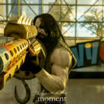 Cosplayer @reallifelobo dressed as Lobo from DC Comics at New York Comic Con 2025, aiming a massive weathered sci-fi blaster toward the camera, wearing white body paint, black facial markings, and a denim vest in front of the NYCC sign inside the Javits Center.