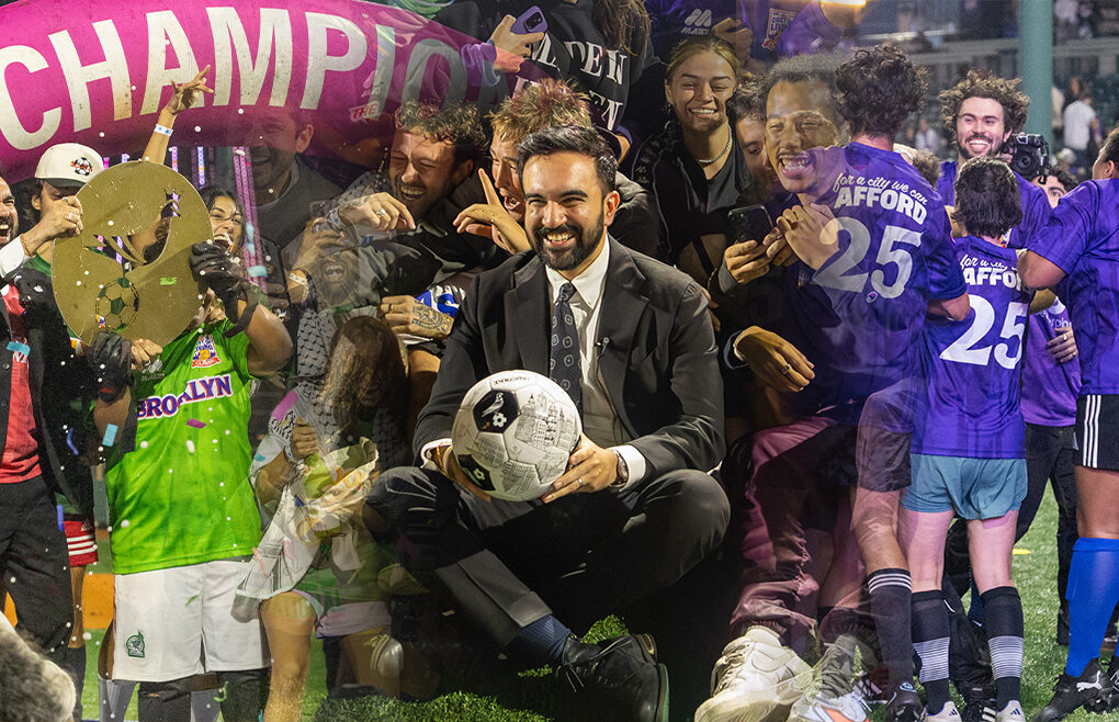 Zohran Mamdani sits smiling with a soccer ball surrounded by cheering players and supporters wearing purple and green jerseys at Maimonides Park in Coney Island, under a bright pink “CHAMPIONS” arch during The Cost of Living Classic tournament on October 19, 2025.