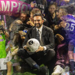 Zohran Mamdani sits smiling with a soccer ball surrounded by cheering players and supporters wearing purple and green jerseys at Maimonides Park in Coney Island, under a bright pink “CHAMPIONS” arch during The Cost of Living Classic tournament on October 19, 2025.