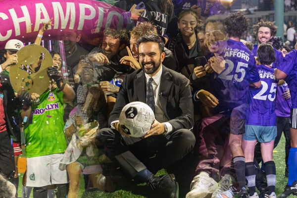 The Cost of Living Classic — Zohran Mamdani Hosts Borough vs. Borough Soccer Tournament in Coney Island Zohran Mamdani sits smiling with a soccer ball surrounded by cheering players and supporters wearing purple and green jerseys at Maimonides Park in Coney Island, under a bright pink “CHAMPIONS” arch during The Cost of Living Classic tournament on October 19, 2025.