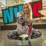 Cosplayer @oliviabettyrain dressed as Anti-Venom Spider-Gwen at New York Comic Con 2025, crouching in a web-slinging pose, wearing a black and white symbiote suit with red and teal details, in front of the NYCC sign at the Javits Center.