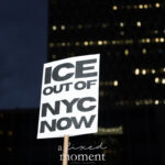 A white protest sign with bold black letters reading “ICE OUT OF NYC NOW” held aloft against the illuminated night skyline of New York City during the ICE OUT of NYC protest on October 22, 2025.