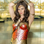 Cosplayer @daphnedaphnedaph dressed as Wonder Woman at New York Comic Con 2025, wearing red and gold armor, silver bracers, and a golden tiara, posing with her arms raised and crossed above her head inside the Javits Center.