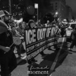 A black-and-white image of protesters marching at night with a large banner reading “ICE OUT OF NYC – STOP THE DEPORTATIONS NOW – ICE FUERA DE NYC! NO MÁS DEPORTACIONES!” during the ICE OUT of NYC protest on October 22, 2025.