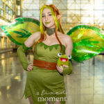 Cosplayer @kingrowletx dressed as a forest fairy at New York Comic Con 2025, wearing a green dress with leaf edges, iridescent green wings, and holding a plush frog, posing inside the Javits Center with sunlight reflecting on the floor.