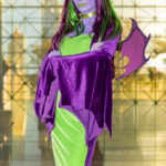 Cosplayer @abbyandrew.yt dressed as a dragon-inspired character at New York Comic Con 2025, wearing a green and purple velvet gown with matching wings, purple skin tone, and green highlights in her hair inside the Javits Center.