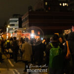 A crowd of protesters march down a city street at night holding signs that say “ICE OUT OF NYC NOW” and “ICE FUERA AHORA” during the ICE OUT of NYC protest in Manhattan on October 22, 2025.