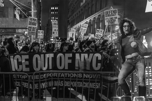 A protester stands above a large crowd holding a banner reading “ICE OUT OF NYC” during a nighttime march from Foley Square to 26 Federal Plaza in New York City, following ICE raids on Canal Street on October 22, 2025.