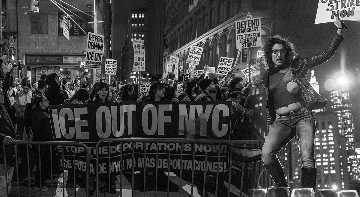A protester stands above a large crowd holding a banner reading “ICE OUT OF NYC” during a nighttime march from Foley Square to 26 Federal Plaza in New York City, following ICE raids on Canal Street on October 22, 2025.
